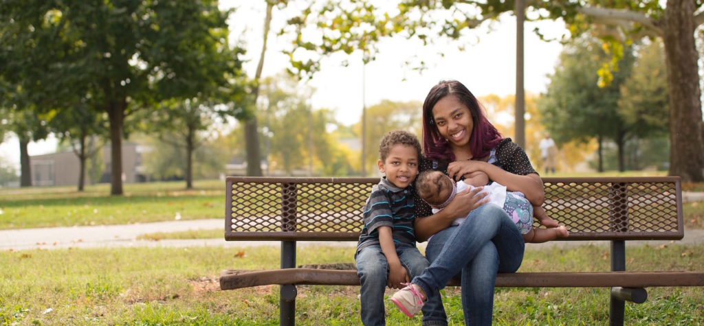 Woman sitting on a bench, nursing a baby with young child sitting next to her