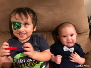 Two children sitting on sofa. Older child holding red and blue plastic toy parts and looking up, infant looking at camera.