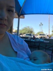 Woman sitting at a table, looking unsmilingly at camera, breastfeeding an infant.