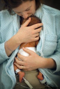 Mother is blue bathrobe holding and kissing a small baby in diaper, skin to skin. IV tubes and monitor tubes for the baby are showing. 