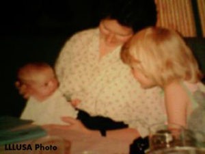 Woman is holding on her lap an infant wearing a cute and comfy pink and white top (or dress) and showing the infant to an older child.