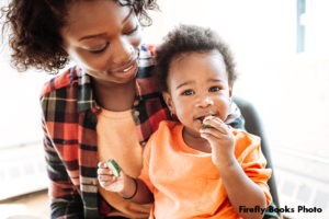 Mother and toddler are sitting together, with the mother looking at the toddler and the toddler looking at the camera while eating a piece of vegetable.