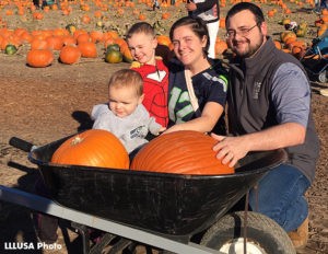 Family kneeling in front of a wheel barrow full of pumpkins.