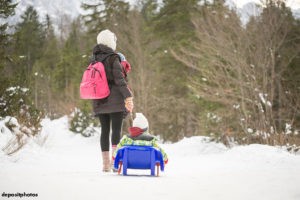 Mother walking in the snow, carrying a backback, a baby and pulling a sled with a toddler