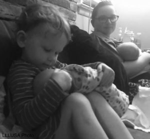 Kylie nursing her son while her daughter nurses her doll sitting on couch
